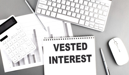 Notebook with the words VESTED INTEREST on a desk with a keyboard, calculator, pen, and charts