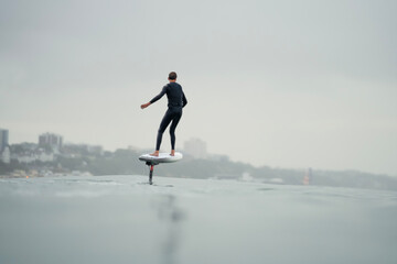 A young man in a wetsuit rides in the sea above the water on an electric powered hydrofoil board, efoil, e-foil, foil drive, hybrid foil.