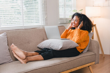 Smiling African American woman relaxing on a cozy couch, wearing headphones, using a laptop, enjoying a comfortable home.