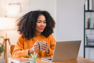 Focused African American woman holding a coffee mug while working on a laptop at home, sitting at a desk with books.