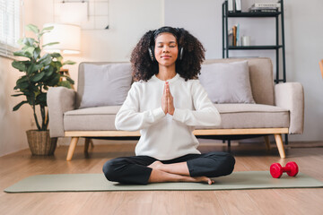 Relaxed African American woman meditating in seated position on yoga mat at home, listening to music with headphones.