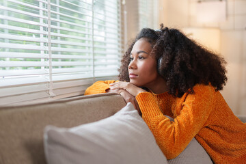 Thoughtful African American woman, resting on a couch, gazing out of a window with a serene expression.