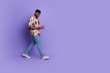 Full length photo of handsome young guy hold device walking dressed stylish colorful garment isolated on purple color background