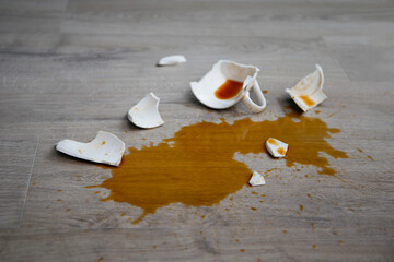 A broken cup of coffee fell on laminate, coffee spilled on floor.