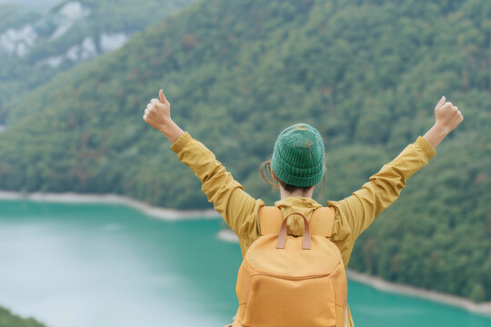 A person with a yellow backpack, arms raised, celebrates in front of a serene mountain lake surrounded by lush greenery on a hike.