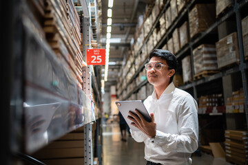 A man wearing glasses is looking at a tablet in a warehouse. He is holding the tablet in his hand and he is focused on the screen. The warehouse is filled with boxes and shelves