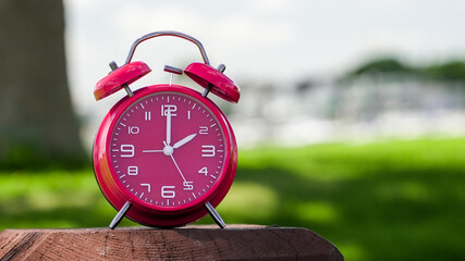 Red alarm clock on wooden surface with blurred green background during daytime showing two o'clock
