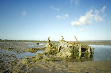 An abandoned half buried rusting and barnacle encrusted 4x4 left to fall apart on a wet sandy beach. A long daytime exposure to blur the water and the clouds.