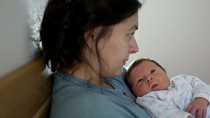 Woman holding her newborn baby in a cozy bedroom, working on a laptop, showcasing the blend of parenting and remote work, modern family life, and nurturing maternal care