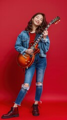 Fototapeta premium A young woman smiles while holding an electric guitar, dressed in stylish casual attire against a vibrant red background