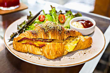 Delicious croissant sandwich filled with ham and cheese, seasoned with herbs, served on ceramic plate with fresh salad and sauce. Perfect for breakfast or brunch