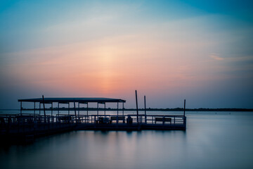 Obraz premium A serene sunset over a tranquil Lewisville lake in The Colony, TX, with a silhouetted pier reflecting soft hues of blue, pink, and orange, creating a peaceful, calming atmosphere