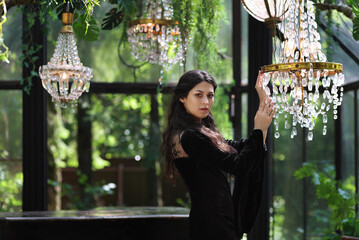 Young brunette woman in black dress standing in glass room full of chandeliers and plants. Fantasy portrait of female adult in evening gown