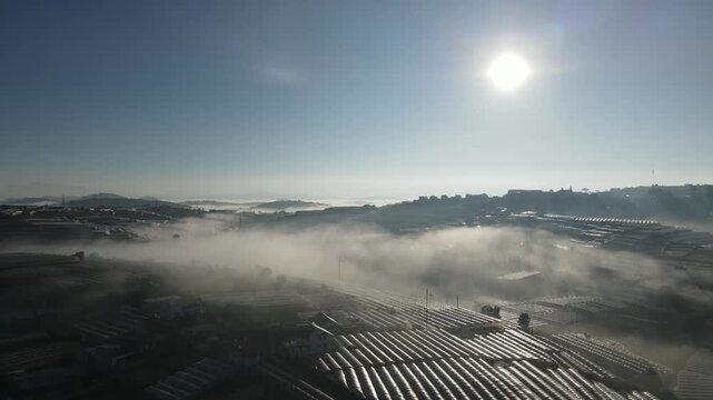 Aerial view of Da Lat city at dawn, nestled amidst rolling hills, with mist enveloping the landscape, and buildings peeping through the fog, showcasing a harmonious blend of nature