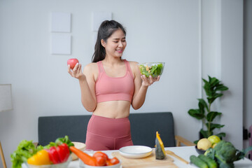 Smiling young woman is holding an apple and a salad bowl, deciding on a healthy meal in her kitchen