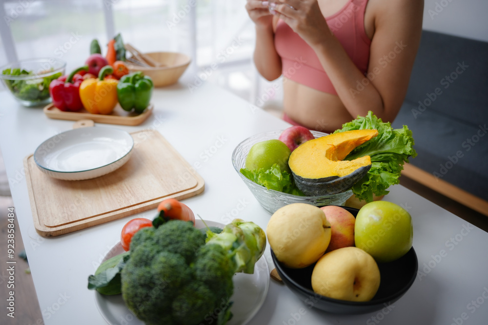 Wall mural Young woman is taking vitamins after preparing a healthy meal full of fruits and vegetables in her kitchen