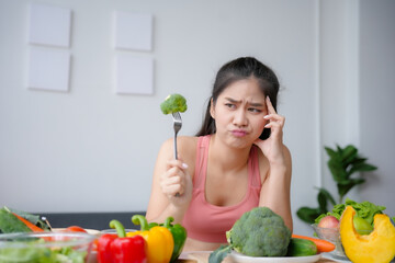 Young woman is holding a fork with a piece of broccoli on it, looking disgusted, with a table full...