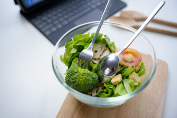 Healthy green salad is served in a glass bowl on a work desk, suggesting a refreshing lunch break