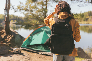 A woman with a backpack looks out over a forest lake. In front of her is a green tent and a beautiful view of peaceful nature at dawn.