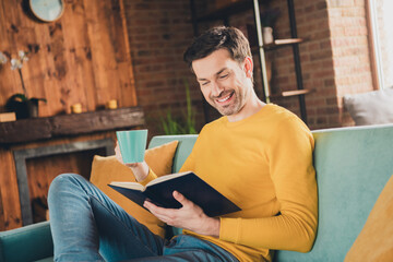 Photo portrait of young guy sit couch read book drink tea dressed yellow garment spend pastime...