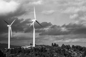 A black and white image depicting two wind turbines on a hilly landscape with a backdrop of mountains and a dramatic cloud-filled sky highlighting the contrast in Tarragona Spain