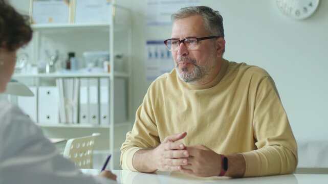 Middle aged male patient describing symptoms of illness to female doctor taking medical records during consultation in clinic
