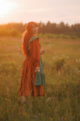 Fototapeta premium A shepherdess woman with red hair and a red and green dress walks on a field of flowers. Rural scene concept with a romantic peasant woman in a linen dress. Romance novel.