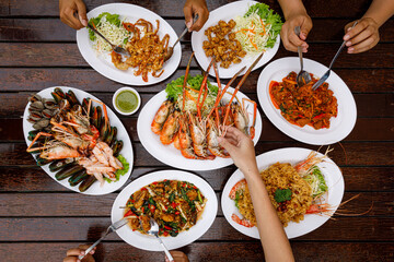 Top view of the dining table. Hands scooping food including grilled river prawns, seafood, fried chicken tendons, stir-fried chili paste, and fried soft-shell crab with garlic.
