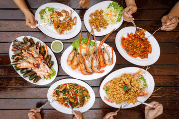 Top view of the dining table. Hands scooping food including grilled river prawns, seafood, fried chicken tendons, stir-fried chili paste, and fried soft-shell crab with garlic.
