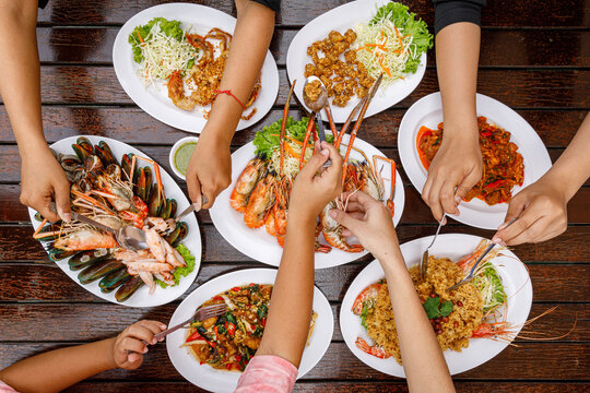 Top view of the dining table. Hands scooping food including grilled river prawns, seafood, fried chicken tendons, stir-fried chili paste, and fried soft-shell crab with garlic.