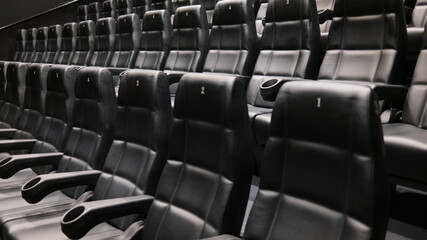Naklejka premium Rows of empty black leather cinema seats with cup holders and numbered backrests in a modern movie theater.