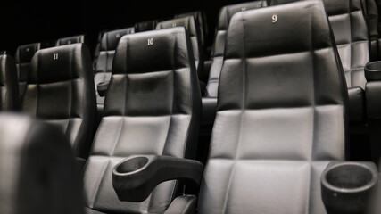 Rows of empty black leather cinema seats with cup holders and numbered backrests in a modern movie...