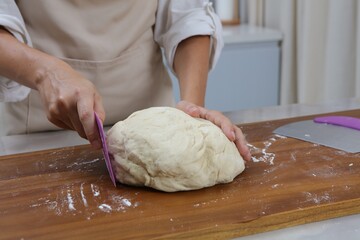 a person wearing a light-colored apron slices a ball of dough on a floured wooden surface. A metal dough scraper lies nearby. The scene, set in a kitchen, captures the hands-on process of preparing