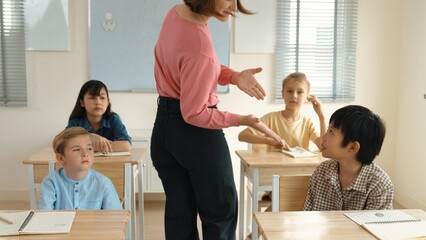 Obraz premium Caucasian teacher walking around classroom while explain classwork. Attraction instructor talking and explain lesson while diverse student listening ans working or taking a note. Education. Pedagogy.