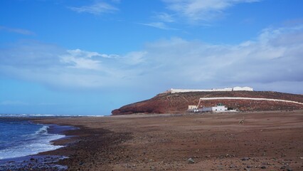 A coastal landscape featuring a rocky beach leading to distant cliffs under a blue sky with scattered clouds, capturing the rugged beauty of the shore of Morocco.
