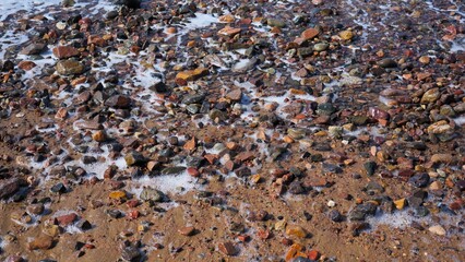 A close-up view of a rocky shoreline featuring various pebbles and stones, with small waves washing over the sand and rocks.
