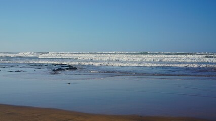 A serene scene of gentle ocean waves lapping against a sandy beach under a clear blue sky, capturing the peacefulness of coastal nature of Morocco.
