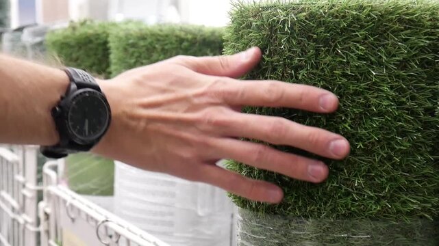 Close-up of many artificial turf rolls on the market and a male hand tries one on the touch