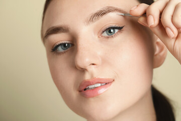 Fototapeta premium Young woman plucking eyebrow with tweezers on beige background, closeup
