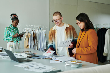 Young male and female designers of attire discussing sketches of new seasonal collection by workplace against colleague with tablet