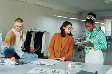 Young brunette female fashion designer looking at screen of tablet held by African American woman during presentation of new items
