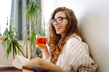 Young woman sitting at home, relaxing in her living room, reading book and drinking tea. Domestic life. Relax and comfort.