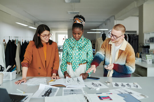 Group of young creative fashion designers discussing new sketches of attire for seasonal fashion collection by workplace