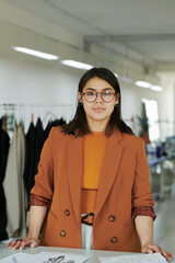 Young confident tailor in smart casualwear looking at camera while standing by workplace with sketches of new attire