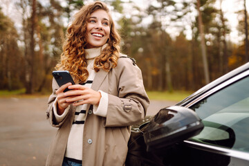 Young woman standing by the car and using phone. Car sharing, rental service or taxi app. Lifestyle, travel, tourism, active life. 