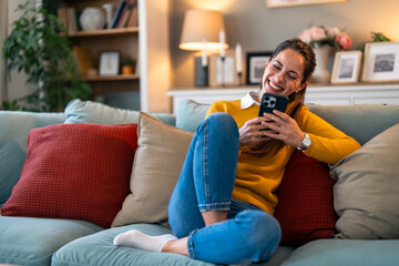 A joyful Caucasian woman interacts with her smartphone, comfortably seated on a couch, surrounded...