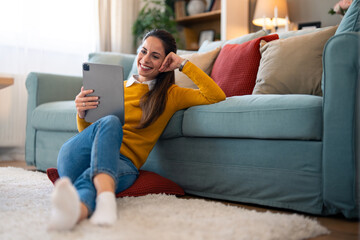 Obraz premium Relaxed, cheerful woman uses a tablet for a video call while sipping coffee, cozily seated on the floor by the couch in her living room.