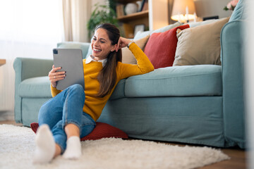A smiling woman sits on the floor, leaning against a couch with a tablet in hand, engaging in a...