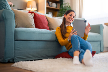 Obraz premium A cheerful caucasian woman enjoys a video chat on her tablet, sipping coffee and sitting casually on the floor by the couch in her cozy living room.
