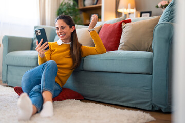 Obraz premium Caucasian woman in casual attire engages in a lively videocall, sitting comfortably on the floor by the couch within a well-lit living room.